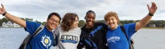 A group of four students in U N E t-shirts pose in front of the ocean off of the Biddeford Campus